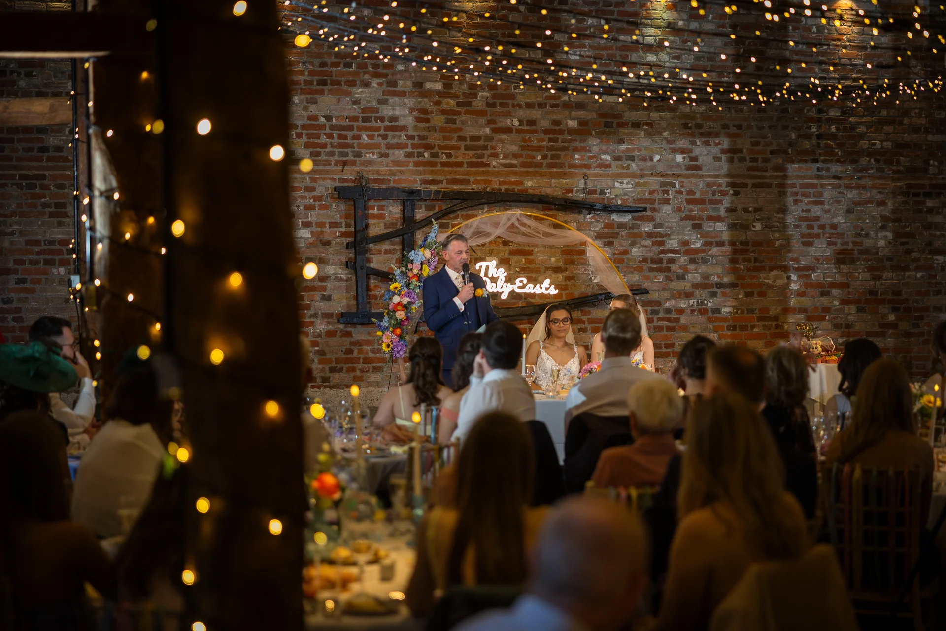 A wedding speech takes place in a rustic barn featuring exposed brick walls and an overhead canopy of sparkling fairy lights.