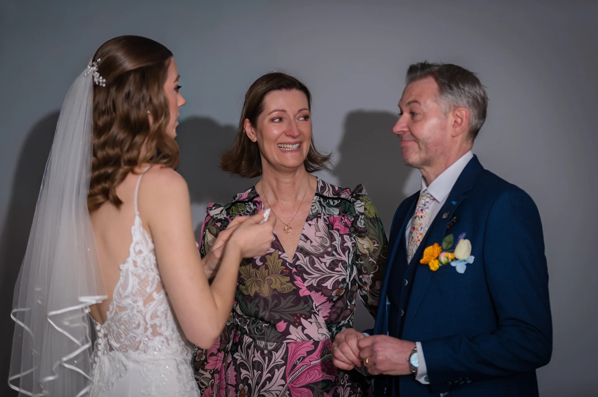 A smiling bride in a lace gown and veil interacts joyfully with her mother in a patterned dress and her father in a blue suit.