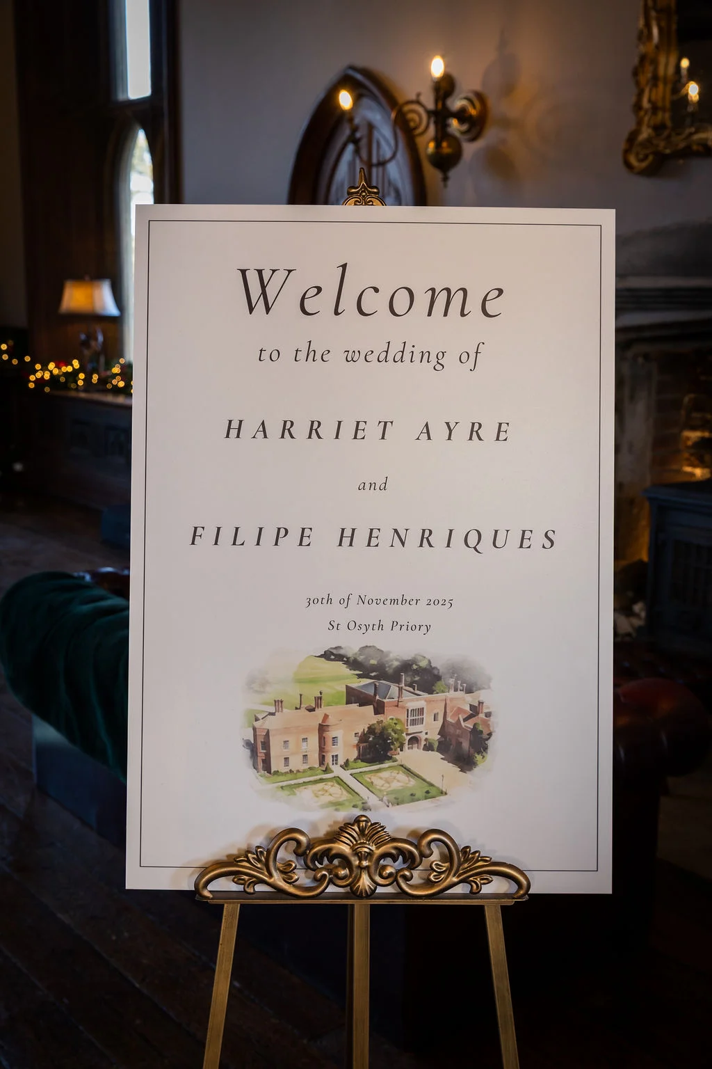An elegant wedding welcome sign featuring a watercolour illustration of the venue sits on an ornate gold easel inside a historic country house.