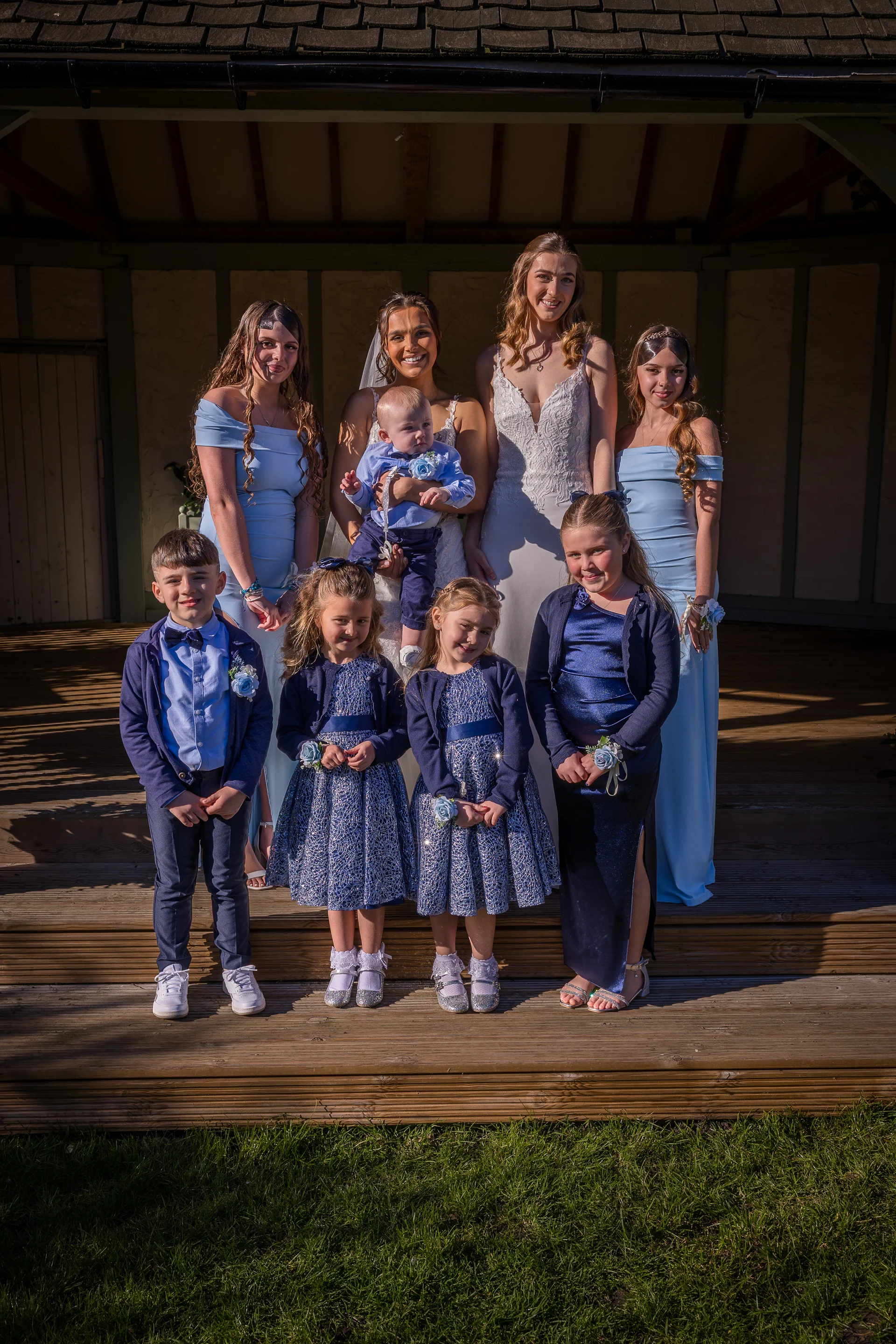 A bride and a group of bridesmaids in dusty blue dresses pose outdoors on wooden steps with young flower girls, a page boy, and a baby.