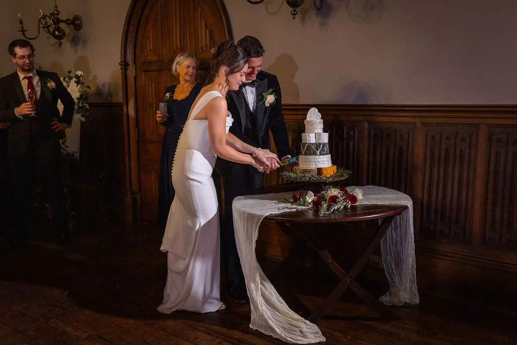 The bride and groom smile together as they cut a multi-tiered cheese wedding cake inside a wood-panelled reception venue, watched by guests.