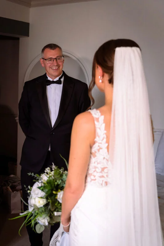 A smiling father in a black tuxedo looks with emotion at his bride daughter wearing a lace gown and veil while holding a bouquet.