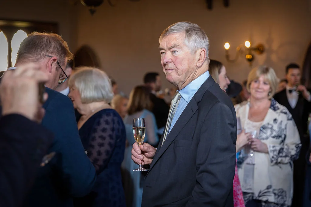 An older male wedding guest holding a glass of champagne during a lively drinks reception with other guests mingling in the background.