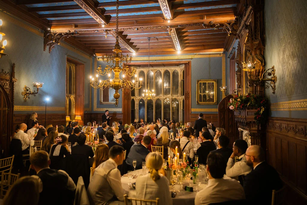 Guests enjoy speeches during a candlelit wedding breakfast inside a grand, wood-panelled stately home ballroom featuring high ceilings and large chandeliers.