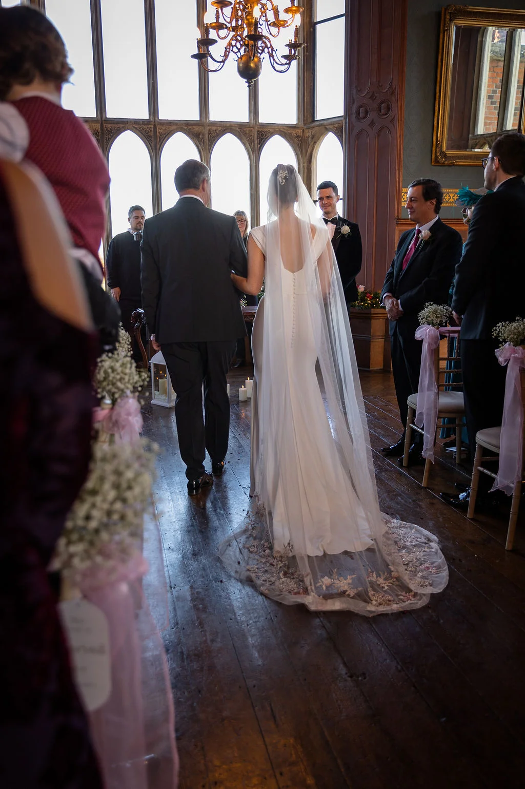 The bride walks down the aisle on her father's arm inside a grand manor house ceremony room featuring tall arched windows and wooden flooring.