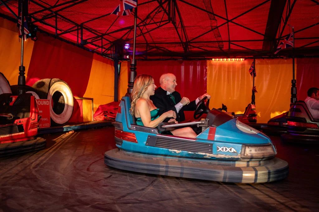 A bride and groom laugh as they drive a blue dodgem car together inside a bright red and yellow funfair tent during their evening wedding reception.