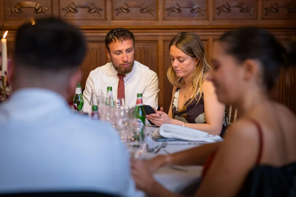 Wedding guests sit around a banquet table in a wood-panelled room, captured candidly through the foreground silhouettes of other attendees.