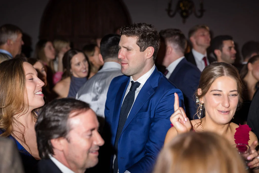 A candid shot of wedding guests mingling and laughing during the evening reception, with a man in a blue suit and dark tie looking towards the side.