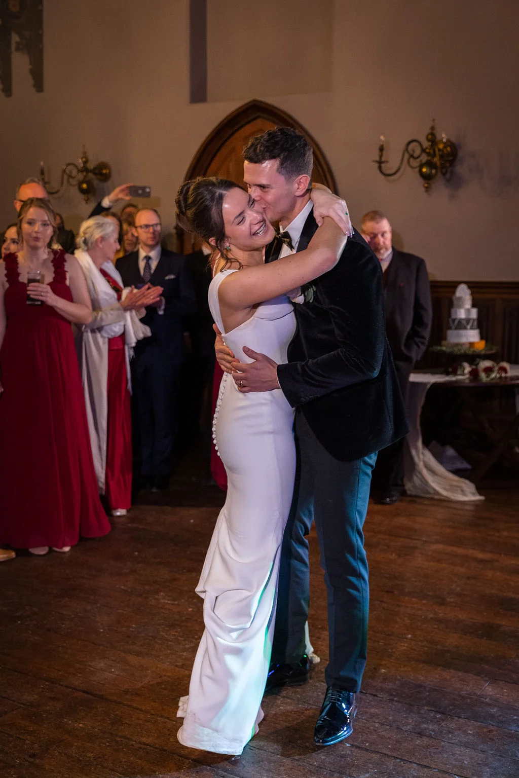 The newly married couple share an emotional and joyful embrace during their first dance, surrounded by admiring wedding guests inside a wood-panelled venue.