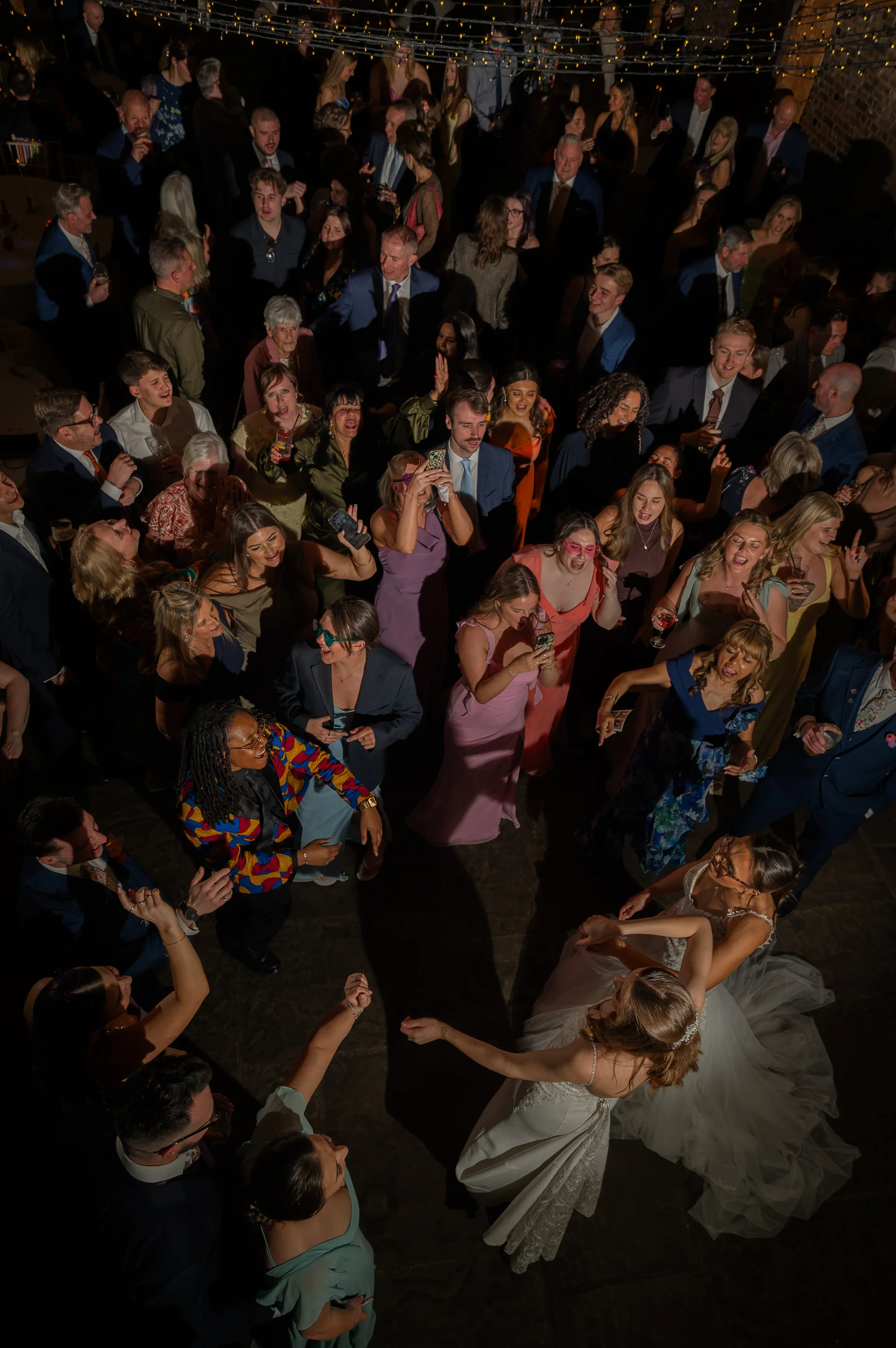 An overhead high-angle shot captures two brides dancing in the centre of a packed, joyful evening wedding reception surrounded by celebrating guests under twinkling fairy lights.