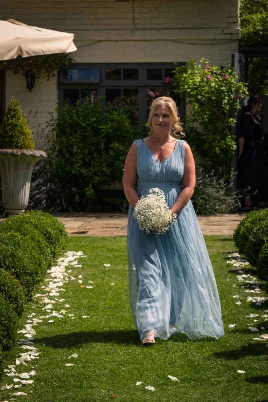 A bridesmaid in a flowing dusty blue chiffon dress walks down a green garden path scattered with white petals, holding a baby's breath bouquet.