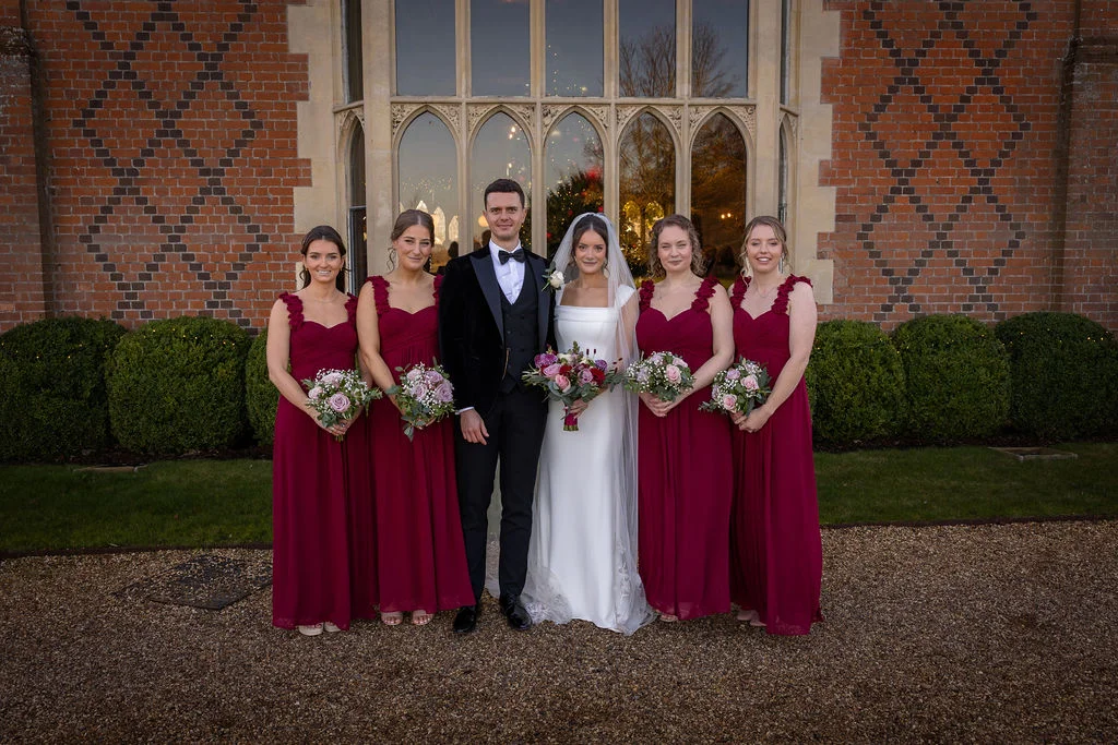 A newlywed couple poses centrally on a gravel driveway in front of a grand red brick manor house with gothic windows, flanked symmetrically by five bridesmaids in elegant burgundy dresses.