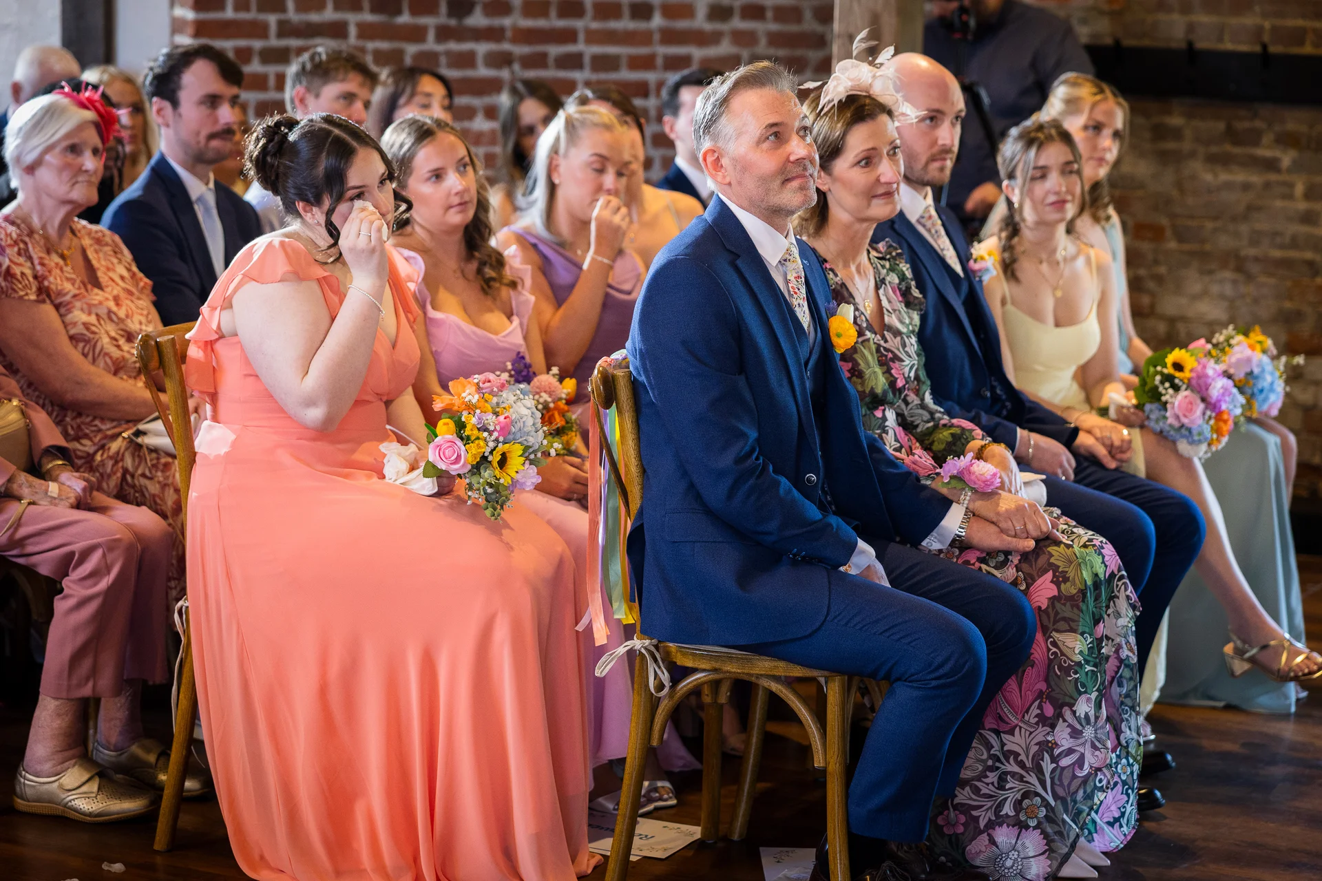 Emotional wedding guests are seated on wooden chairs inside a rustic brick barn venue, with one bridesmaid wiping away tears while holding a colourful bouquet.