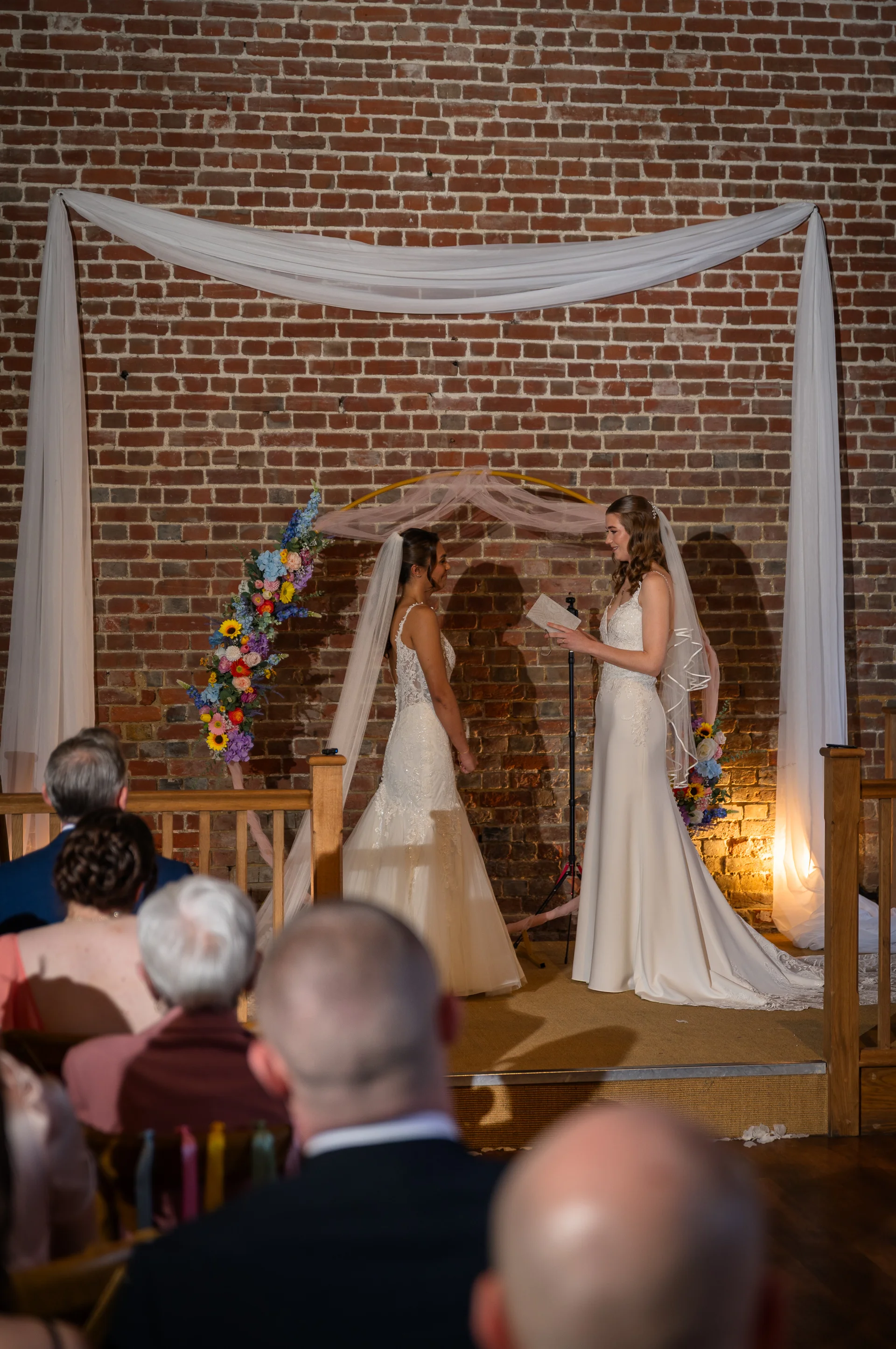 Two brides exchange personal vows during an indoor wedding ceremony against an exposed brick backdrop decorated with a floral arch and white fabric drapes.