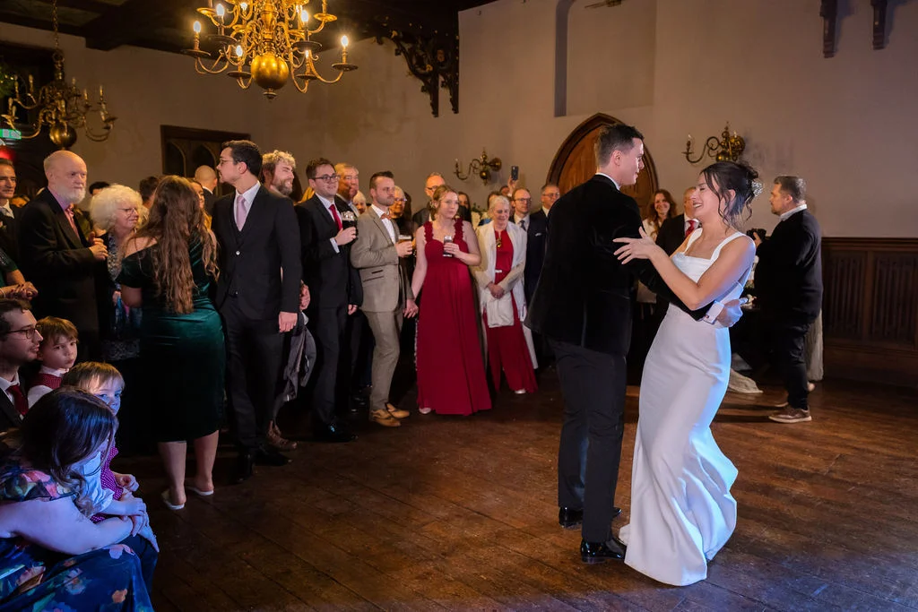 Newlyweds share a joyful first dance on a polished wooden floor inside a historic reception hall, surrounded by smiling guests watching from the sidelines.
