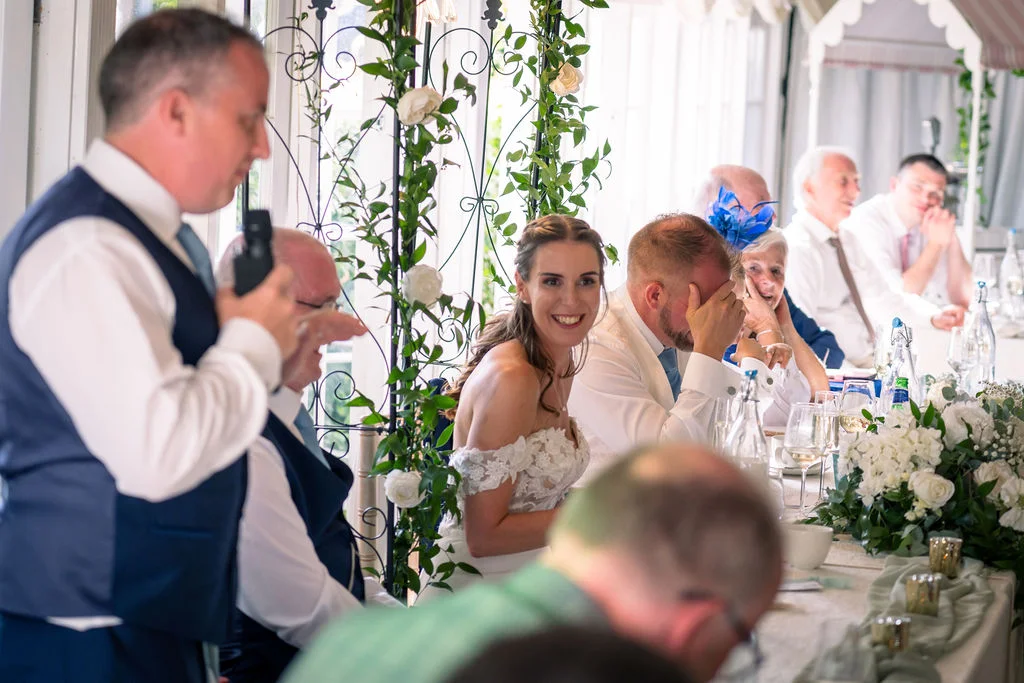 A guest delivers a speech at the top table while the smiling bride and reacting groom listen alongside seated guests.