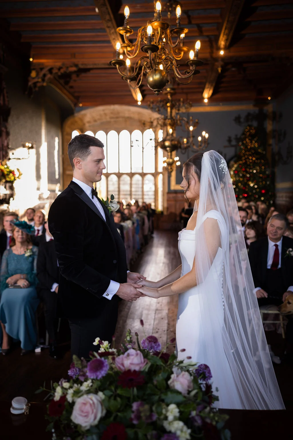 A bride and groom hold hands during their wedding ceremony inside a grand, historic hall lit by magnificent chandeliers and soft window light.