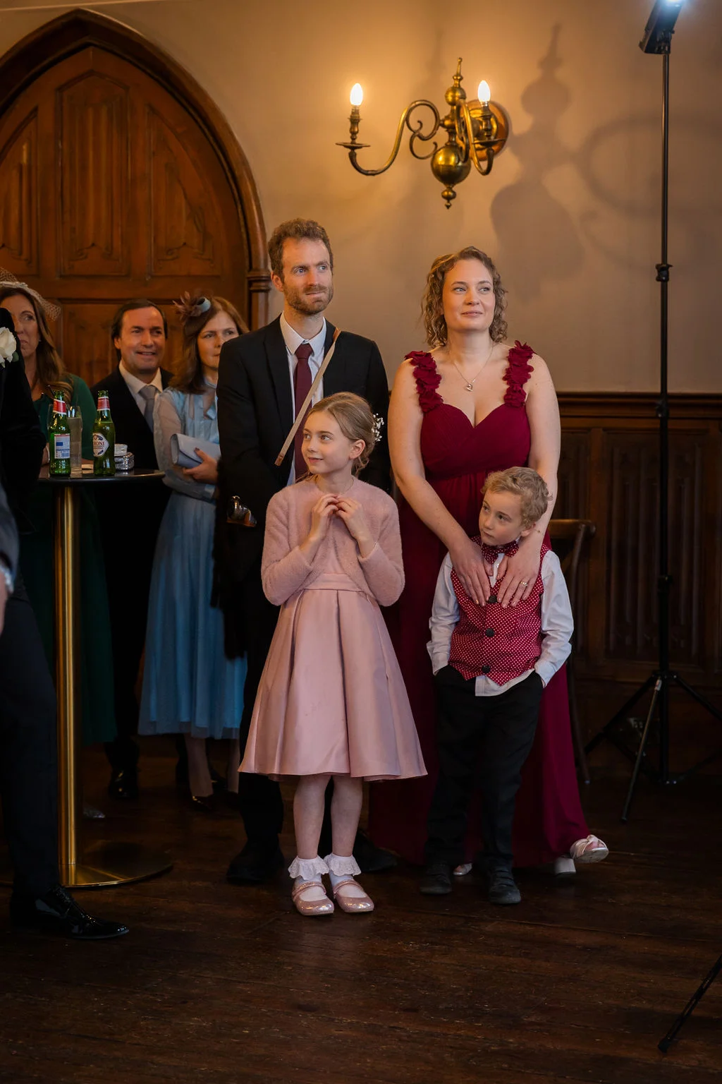 A family and other wedding guests watch happily together inside a rustic venue with wooden floors and traditional panelling.