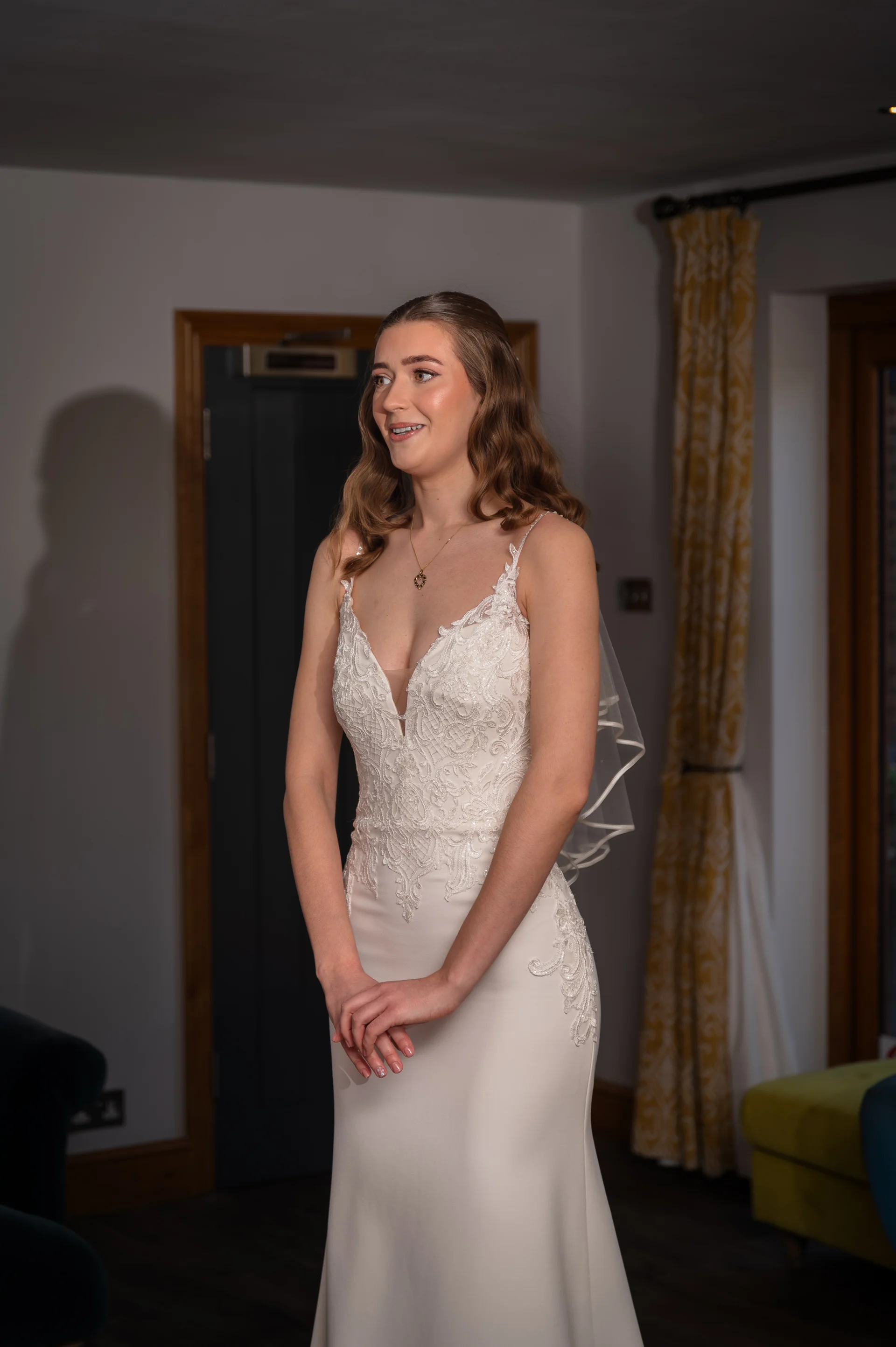 A beautiful bride smiling softly while wearing an elegant V-neck crepe wedding gown and a sheer elbow-length veil during her morning preparations indoors.