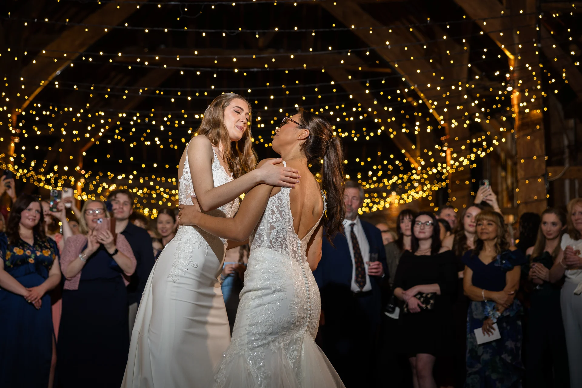 Two brides share a tender first dance beneath a breathtaking canopy of fairy lights inside a rustic barn filled with watching guests.