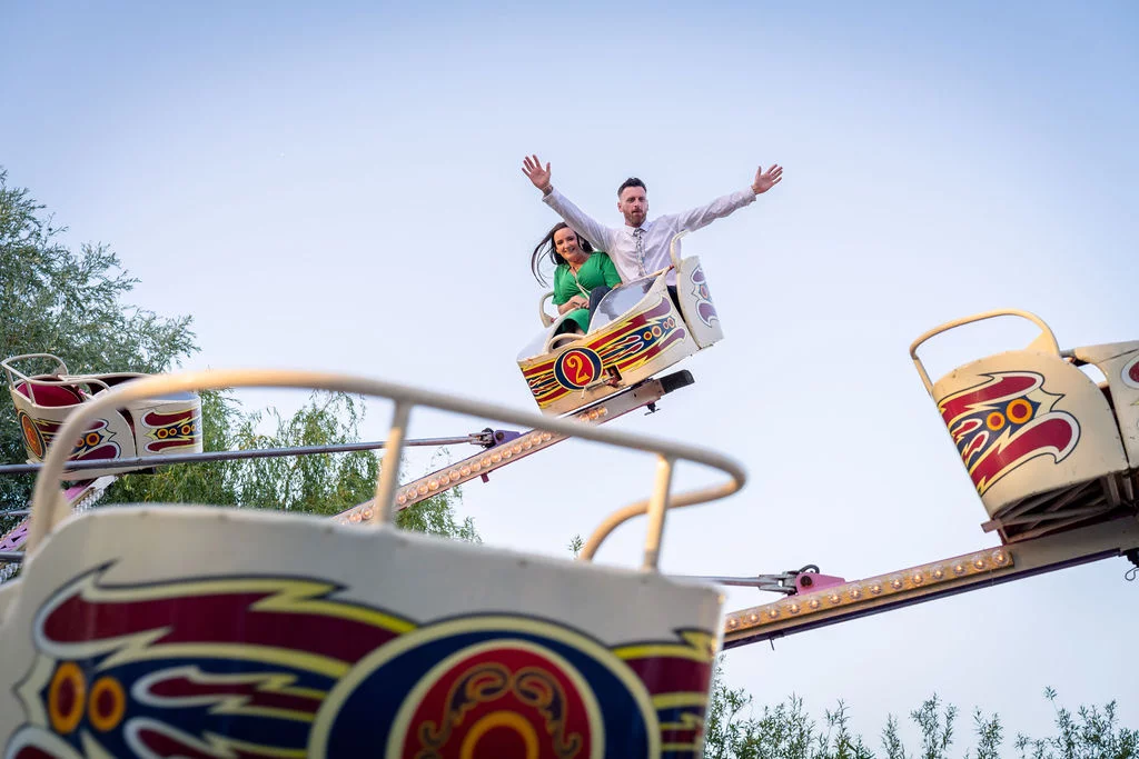 A newlywed couple with arms raised in joy as they ride a vintage funfair car against a clear blue sky.