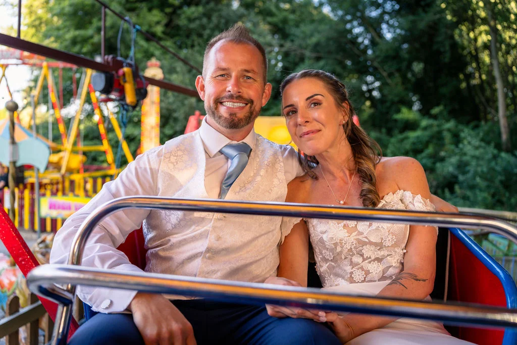 A smiling bride and groom sit together on a colourful funfair ride during their wedding celebrations.