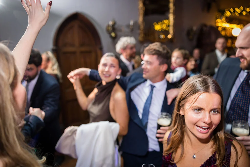 Wedding guests enjoy a lively evening reception, dancing and laughing together in a warmly lit venue with a stone archway in the background.