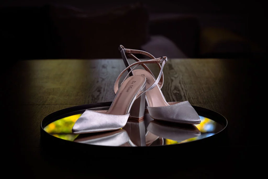 A pair of elegant satin bridal block heel shoes resting on a circular mirror tray, captured in a moody and atmospheric close-up shot.