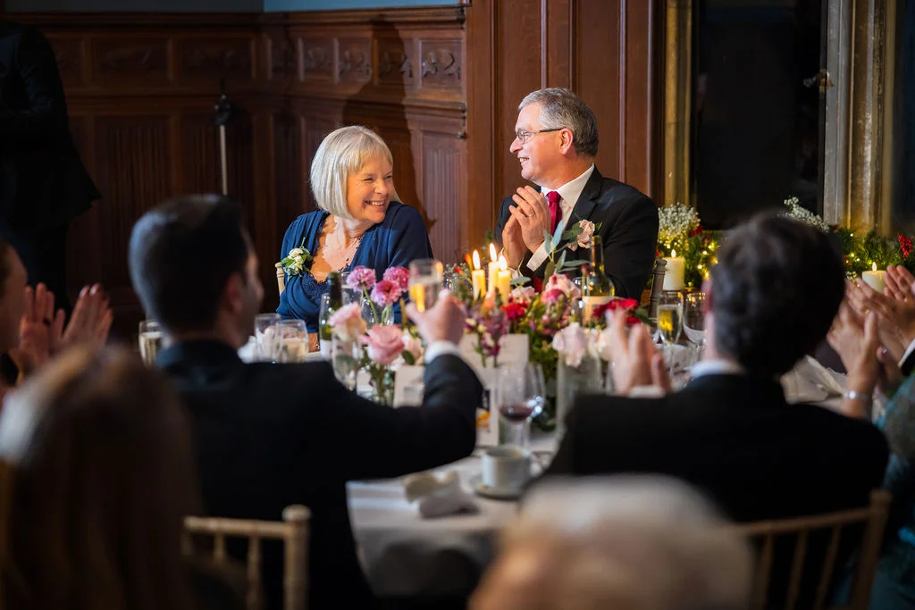Wedding guests applaud and smile as a mature couple seated at the top table share a joyful moment during the reception speeches.
