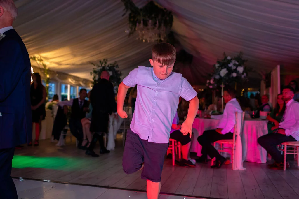 A young boy energetically dances on a illuminated dance floor inside a decorated marquee with soft coloured lighting.