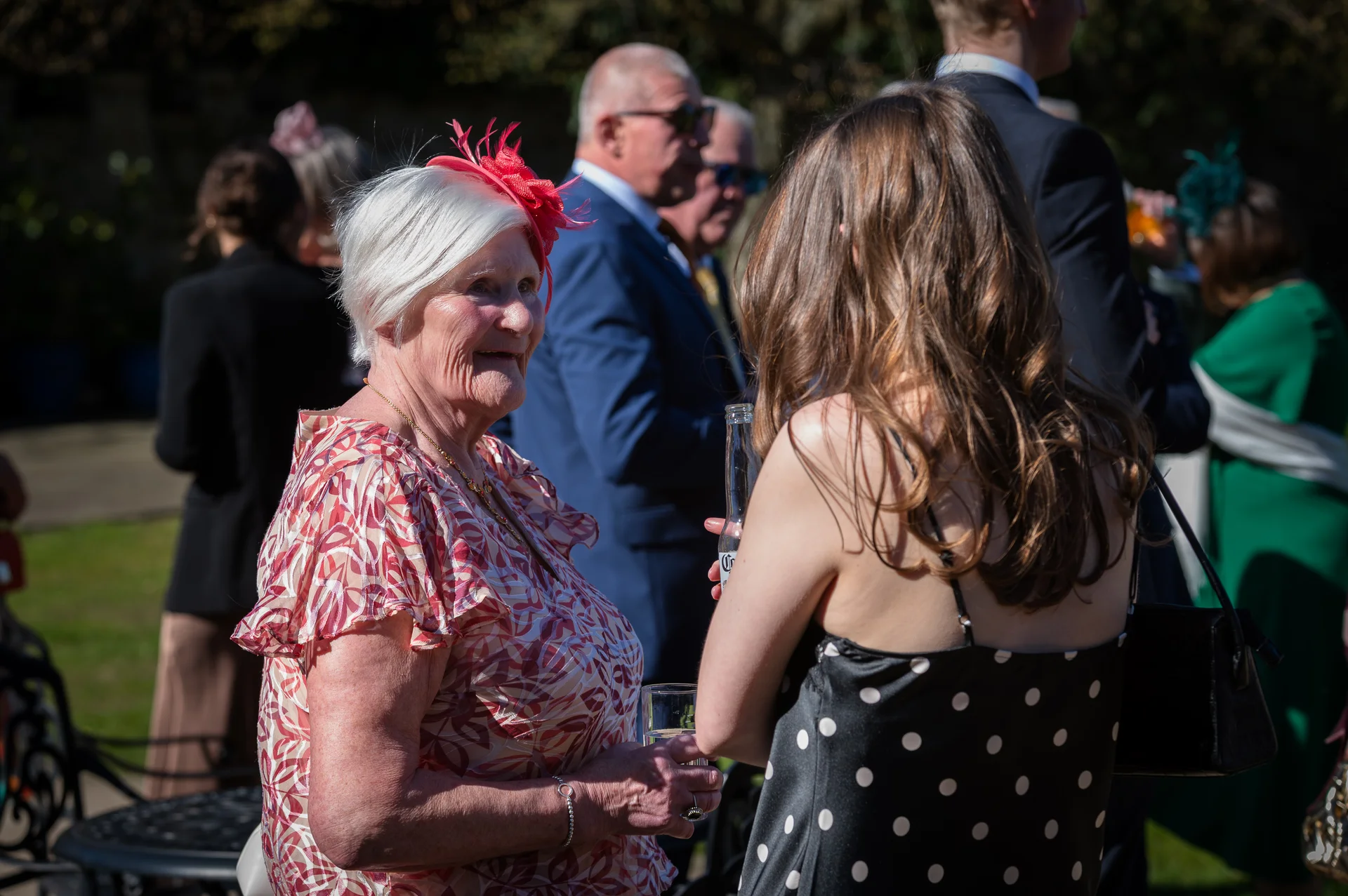 An elderly woman with white hair and a red fascinator chats and laughs with a younger guest during an outdoor wedding drinks reception.