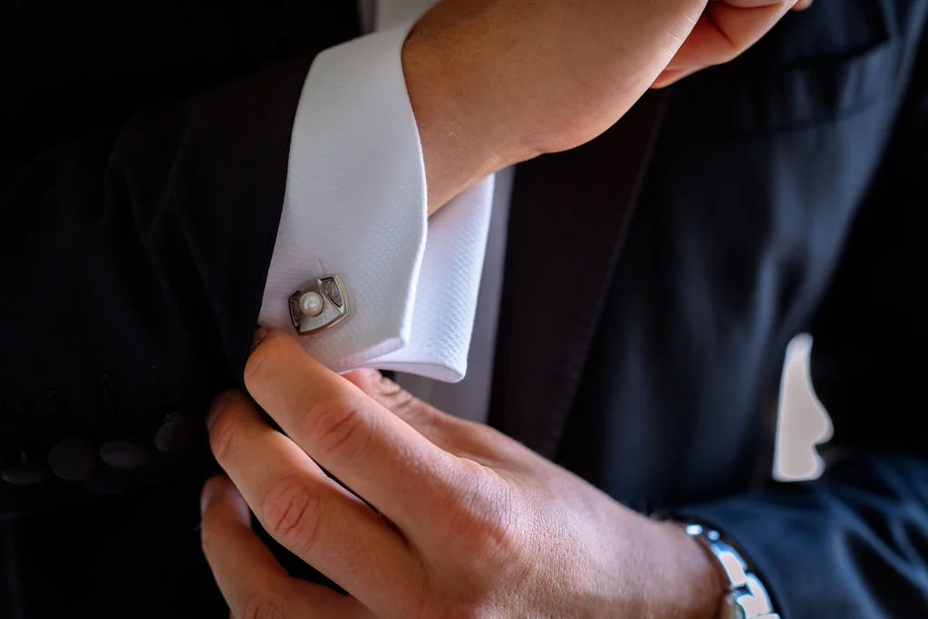 A close-up shot of a groom carefully fastening his distinctive cufflinks on his crisp white shirt and black tuxedo during morning preparations.