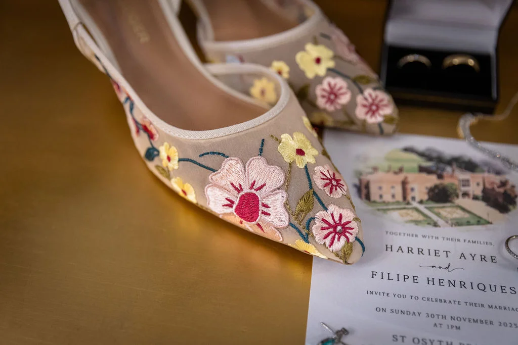 An elegant bridal flat lay featuring embroidered sheer heels alongside a personalised wedding invitation suite and wedding bands in a velvet box.