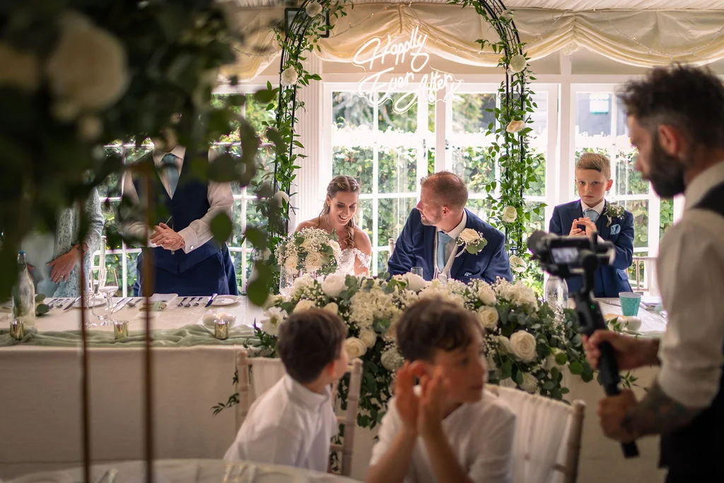The bride and groom sit at the top table during a wedding reception, framed by a floral arch and a glowing neon sign.