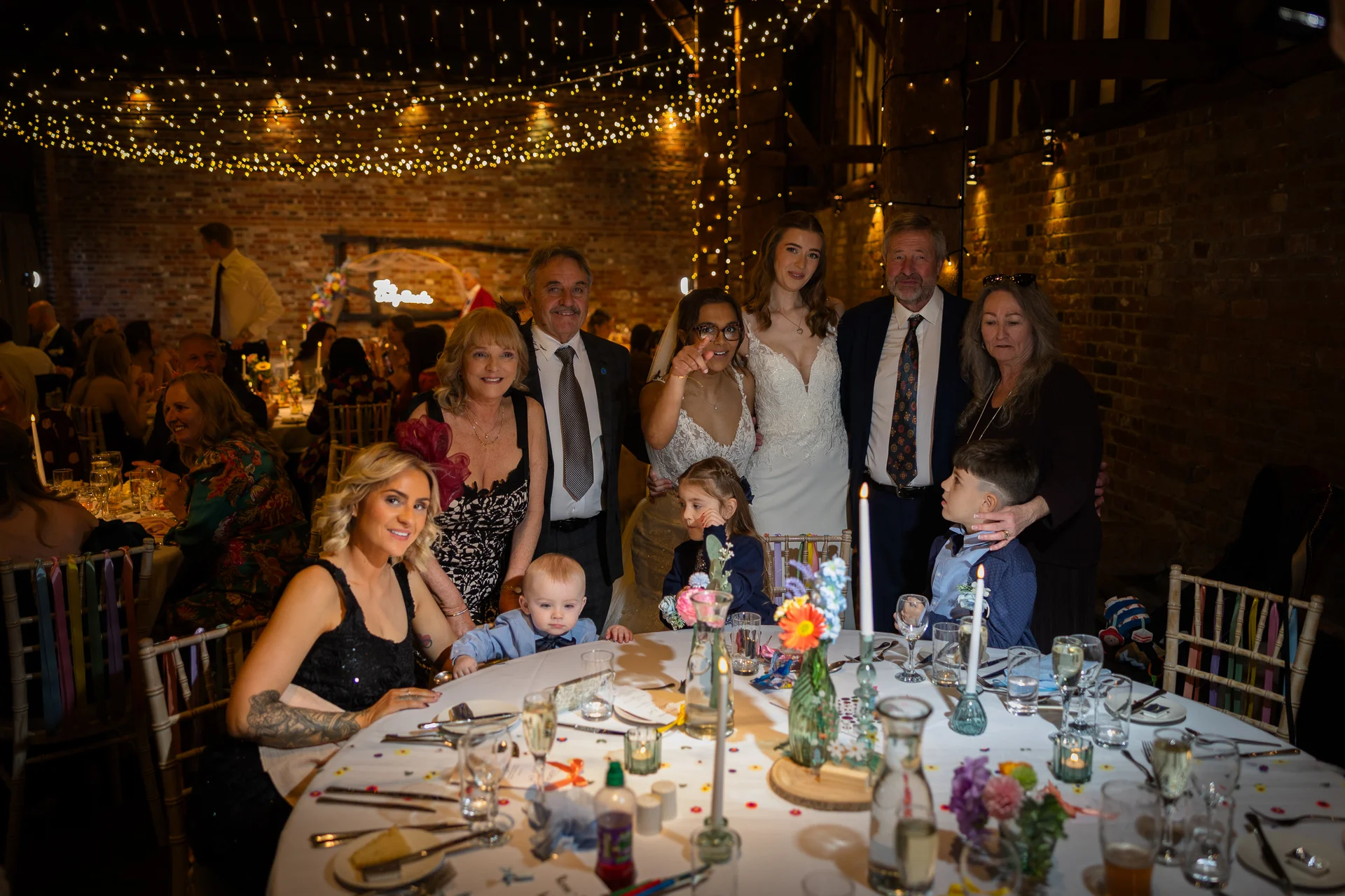 A large family group gathers around a candlelit round dining table at an evening wedding reception inside an atmospheric rustic barn illuminated by a canopy of fairy lights.