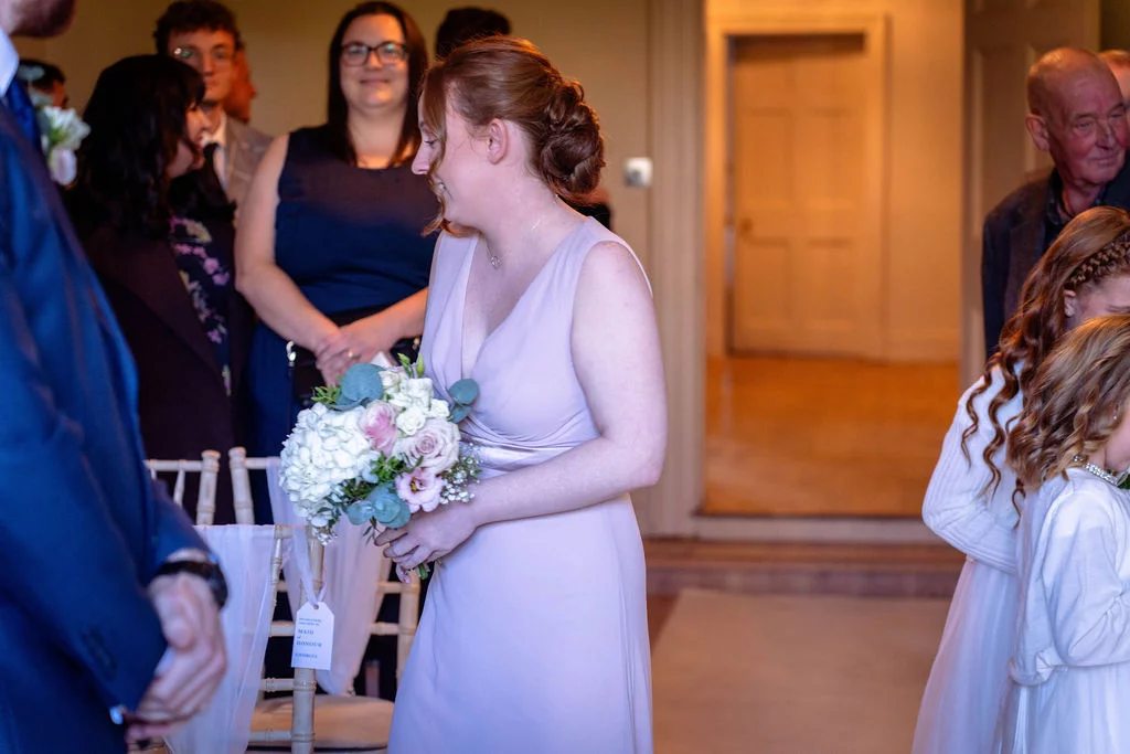 A smiling bridesmaid in a pink dress holding a floral bouquet walks down the aisle during a wedding ceremony.