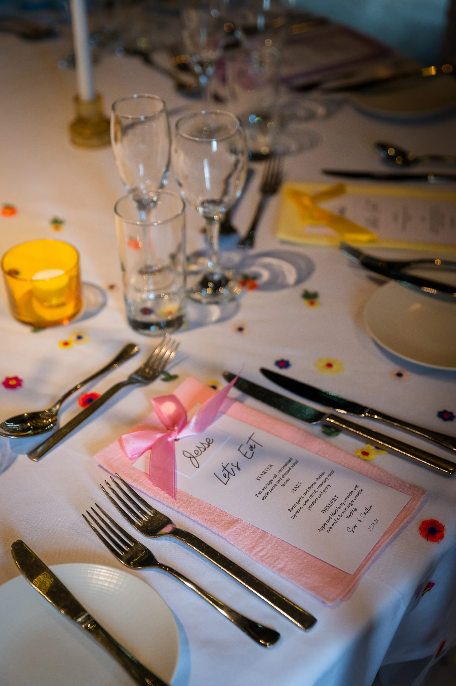 An elegantly styled wedding tablescape features a white embroidered tablecloth, a pink napkin tied with a satin bow, and a printed menu alongside fine cutlery and glassware.