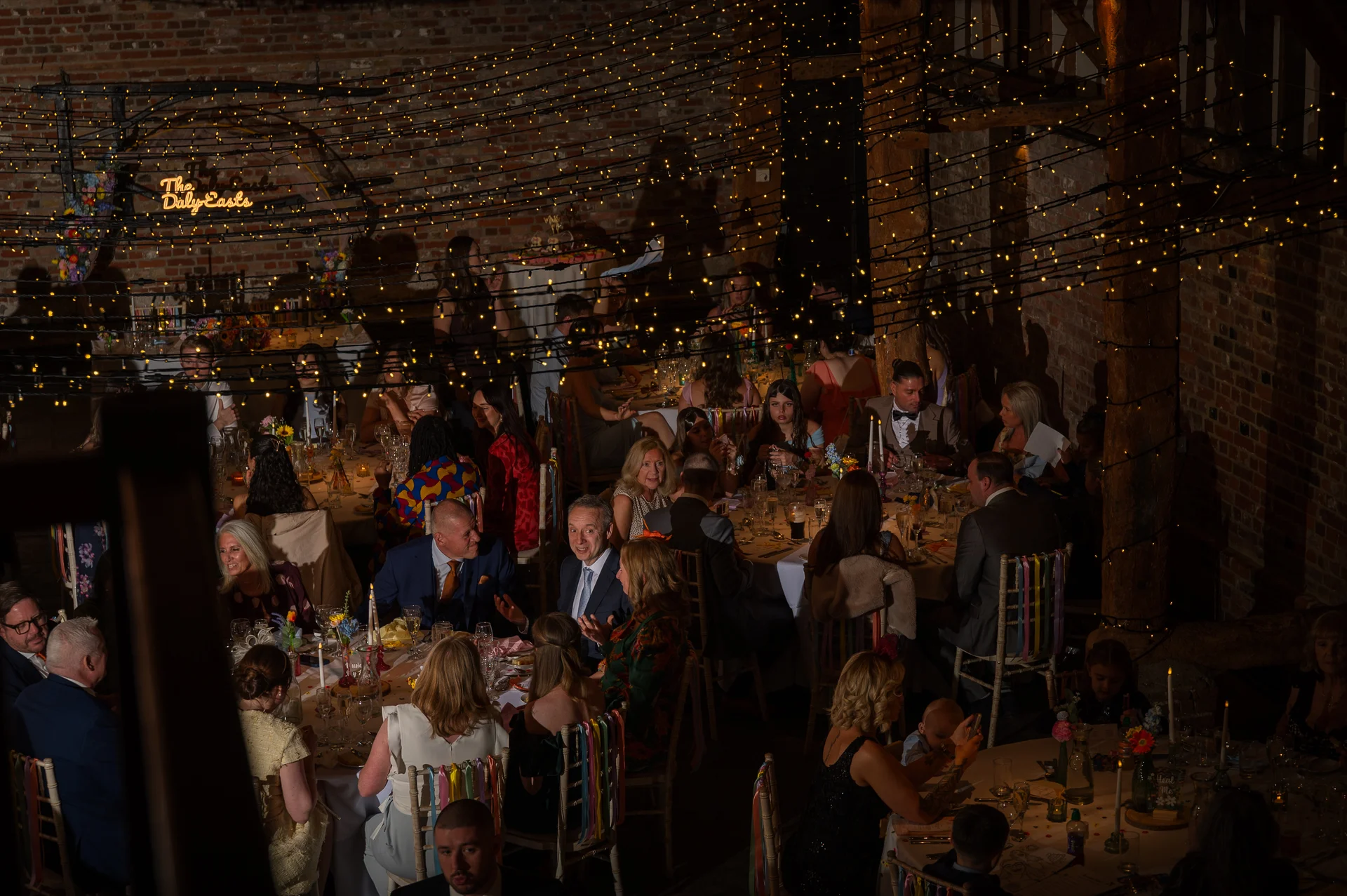 Wedding guests enjoy a candlelit wedding breakfast inside a rustic brick barn illuminated by a vast canopy of twinkling fairy lights.