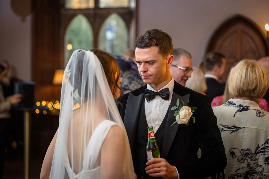 The groom looks lovingly at his bride in her veil while holding a bottled drink during the wedding reception.