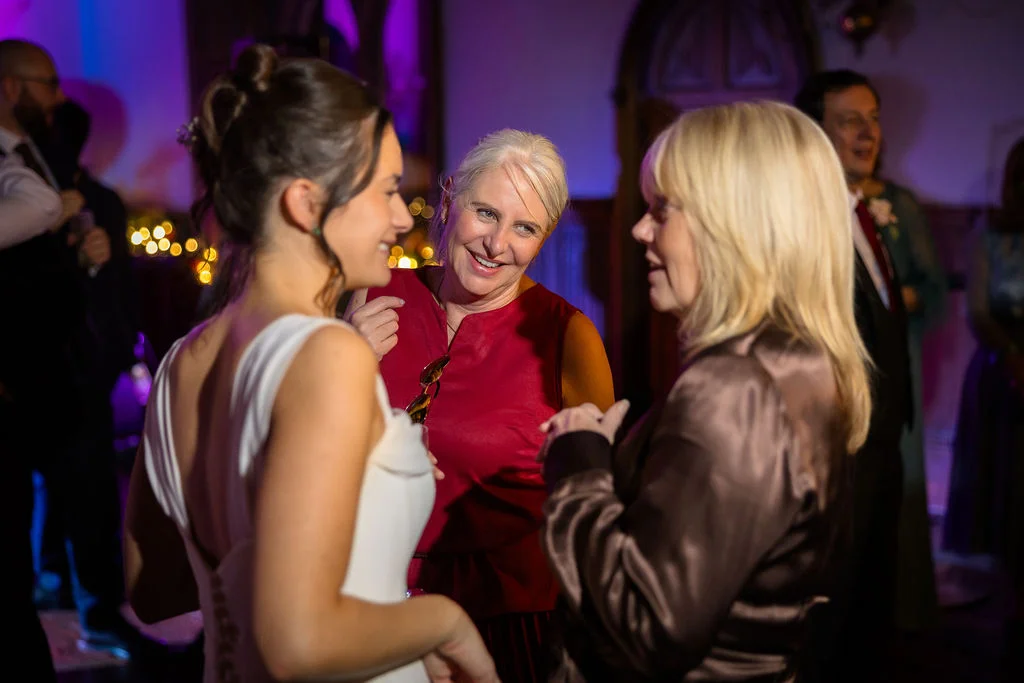A smiling bride in a white wedding dress chats and laughs with guests during an evening reception illuminated by warm purple ambient lighting.