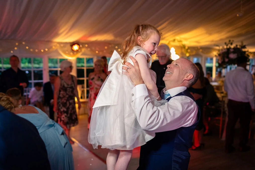 A joyful guest lifts a young flower girl in a white dress high into the air on the dance floor inside a marquee.