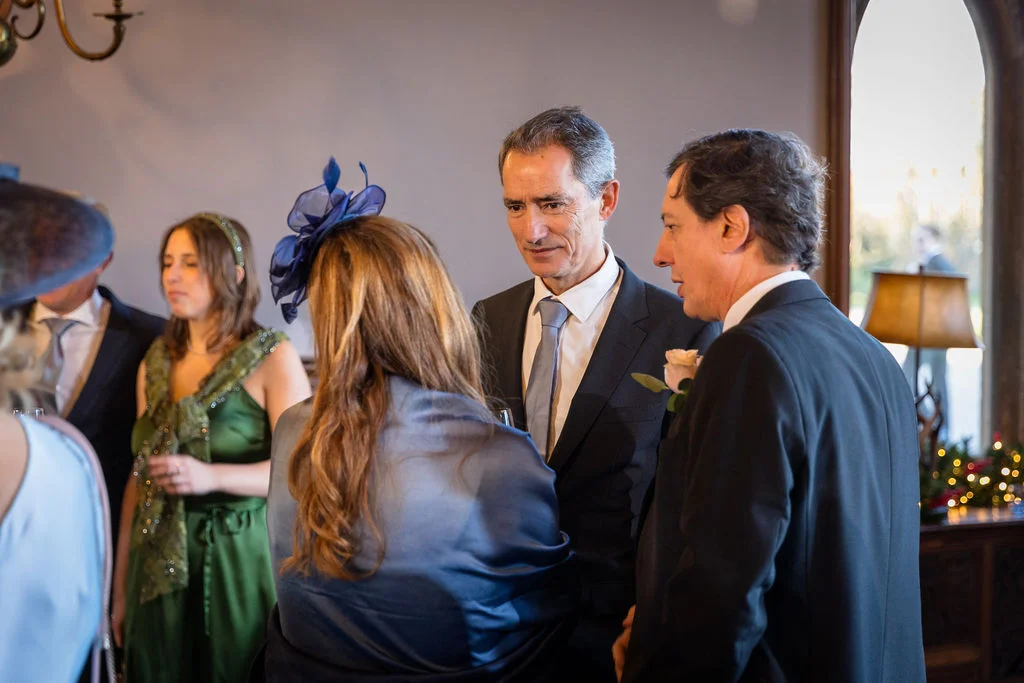Wedding guests in smart suits and a blue fascinator mingle happily together indoors during a reception.