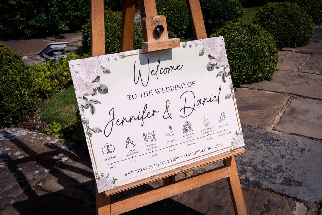 A white welcome sign with floral illustrations and a timeline schedule sits on a wooden easel outdoors in a sunny garden.