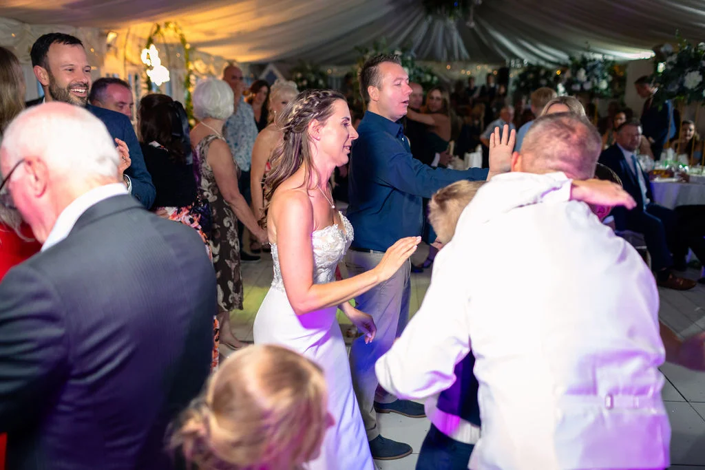 A lively wedding reception in a draped marquee where the bride and guests enjoy dancing together under vibrant party lights.