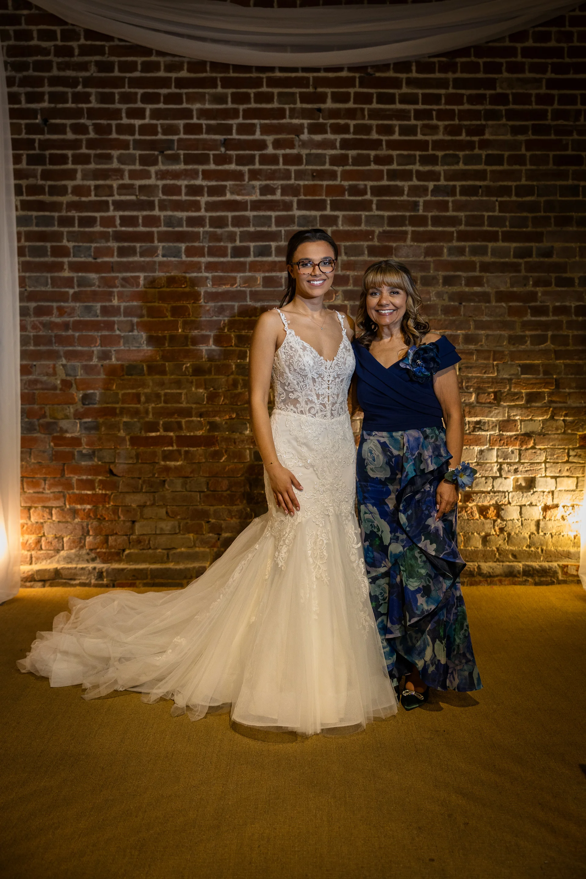 A smiling bride wearing a lace fit and flare gown and glasses poses alongside her mother in a blue patterned dress in front of an exposed brick wall.