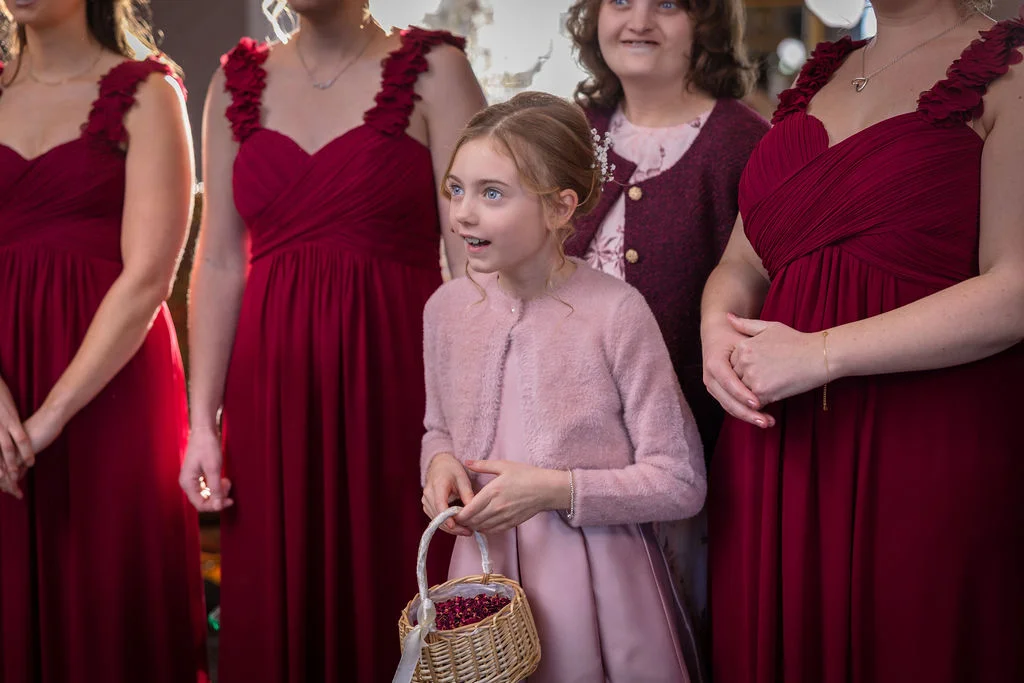 A young flower girl holding a petal basket looks on with a surprised expression, flanked by bridesmaids in matching burgundy dresses.
