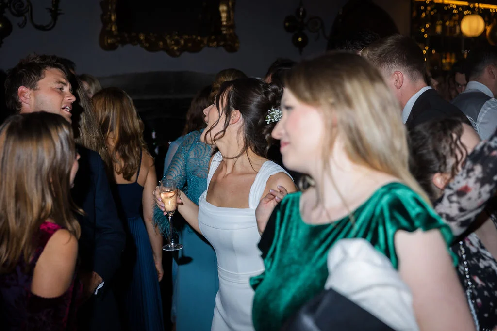 A bride in a white square-neck dress laughs joyfully while holding a glass of champagne on a crowded, lively evening reception dance floor.