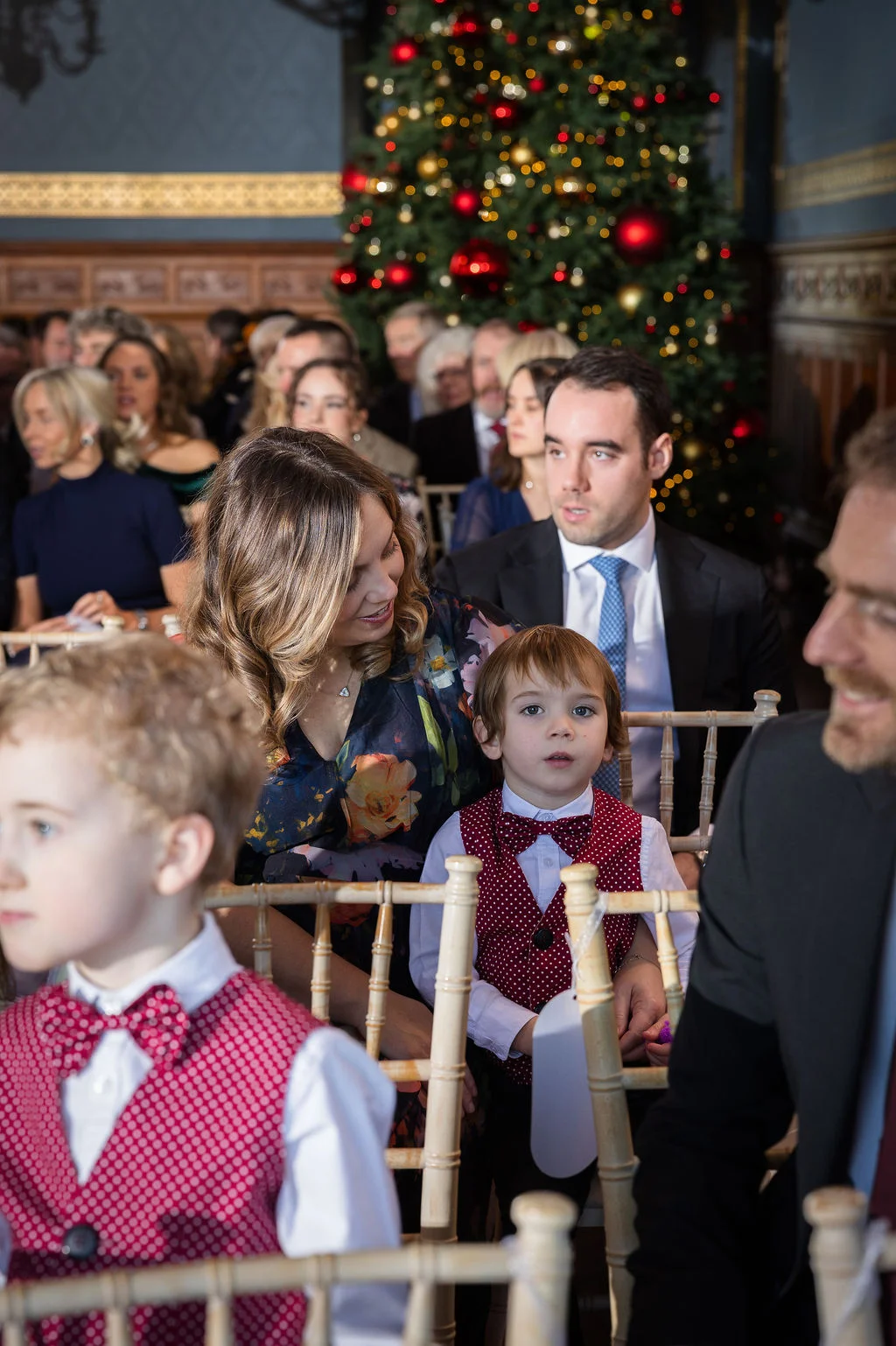 Family and guests seated together inside a venue decorated for a festive Christmas wedding with a decorated tree in the background.
