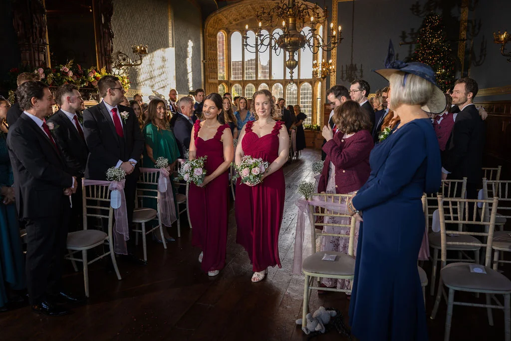 Two bridesmaids in deep burgundy dresses walk down the aisle holding small bouquets during a wedding ceremony inside a grand historic hall with large windows.