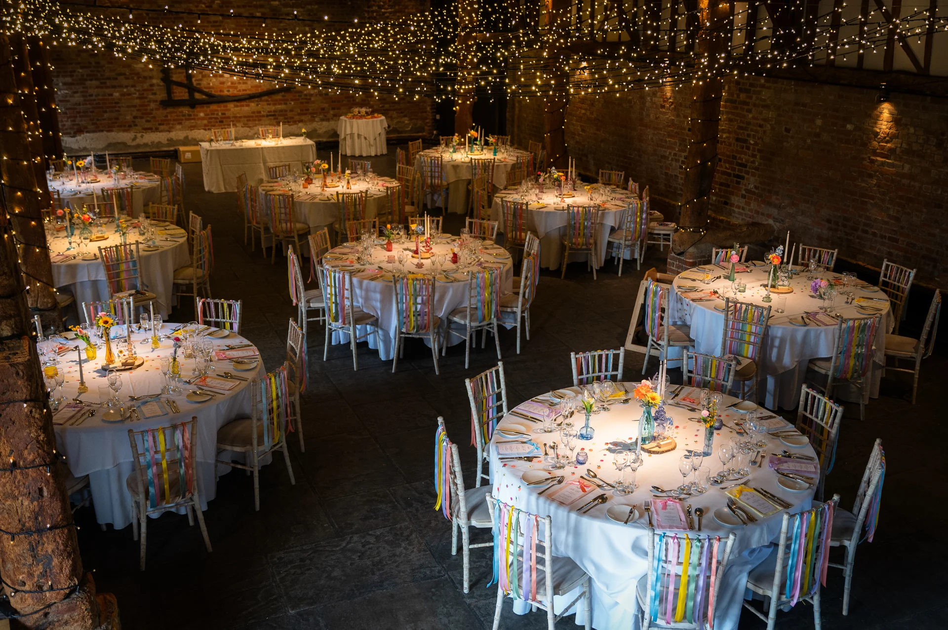 An overhead view of a rustic barn wedding reception featuring round tables adorned with white linens, colourful pastel ribbons on chairs, and a magical fairy light canopy overhead.